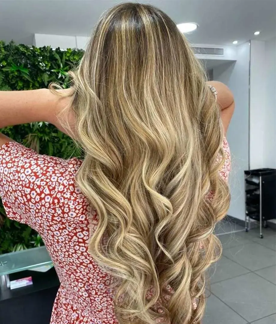 Blonde balayage on long wavy hair, in front of the salon's plant wall